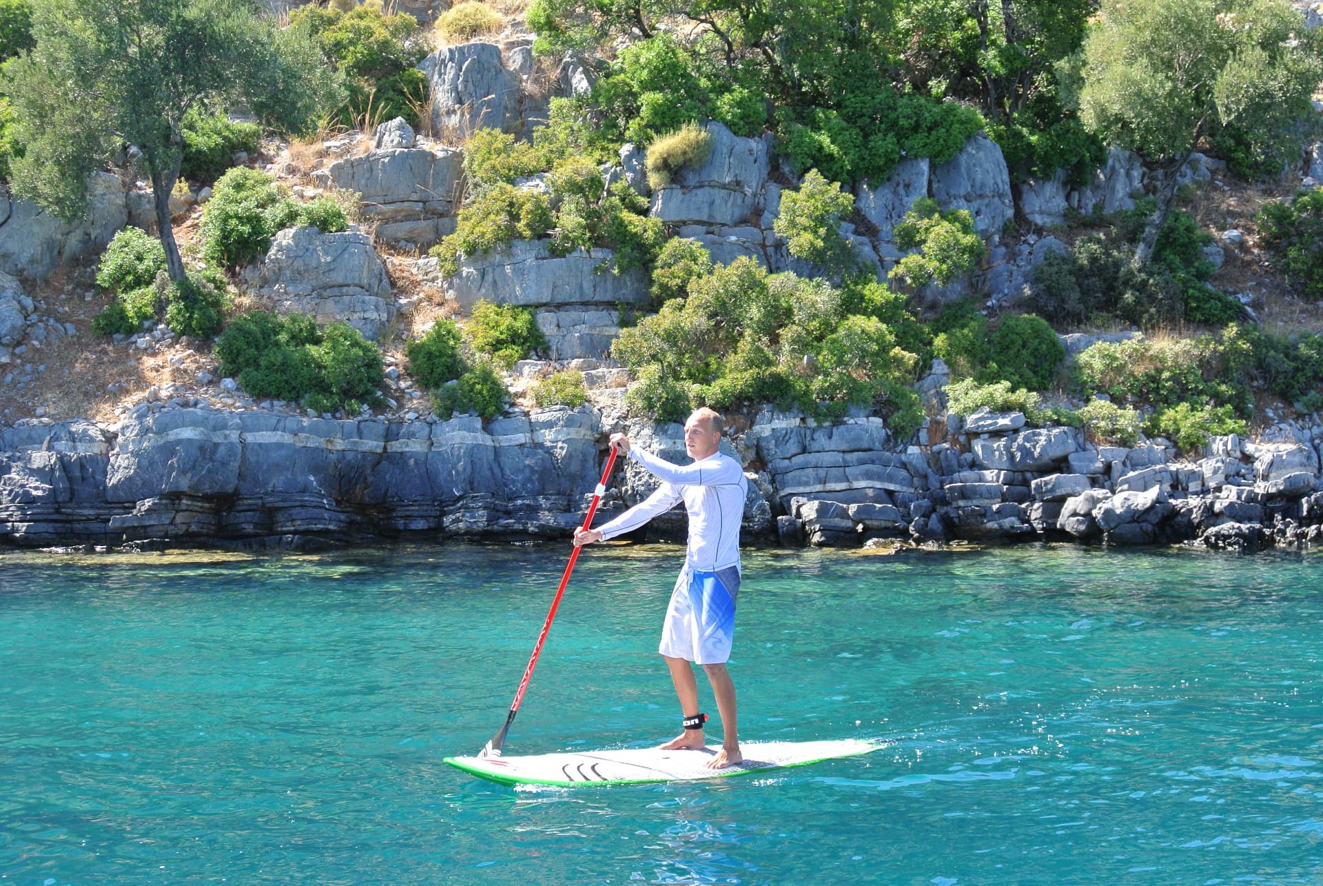 Rickard paddleboarding on clear turquoise water along a rocky Mediterranean shoreline